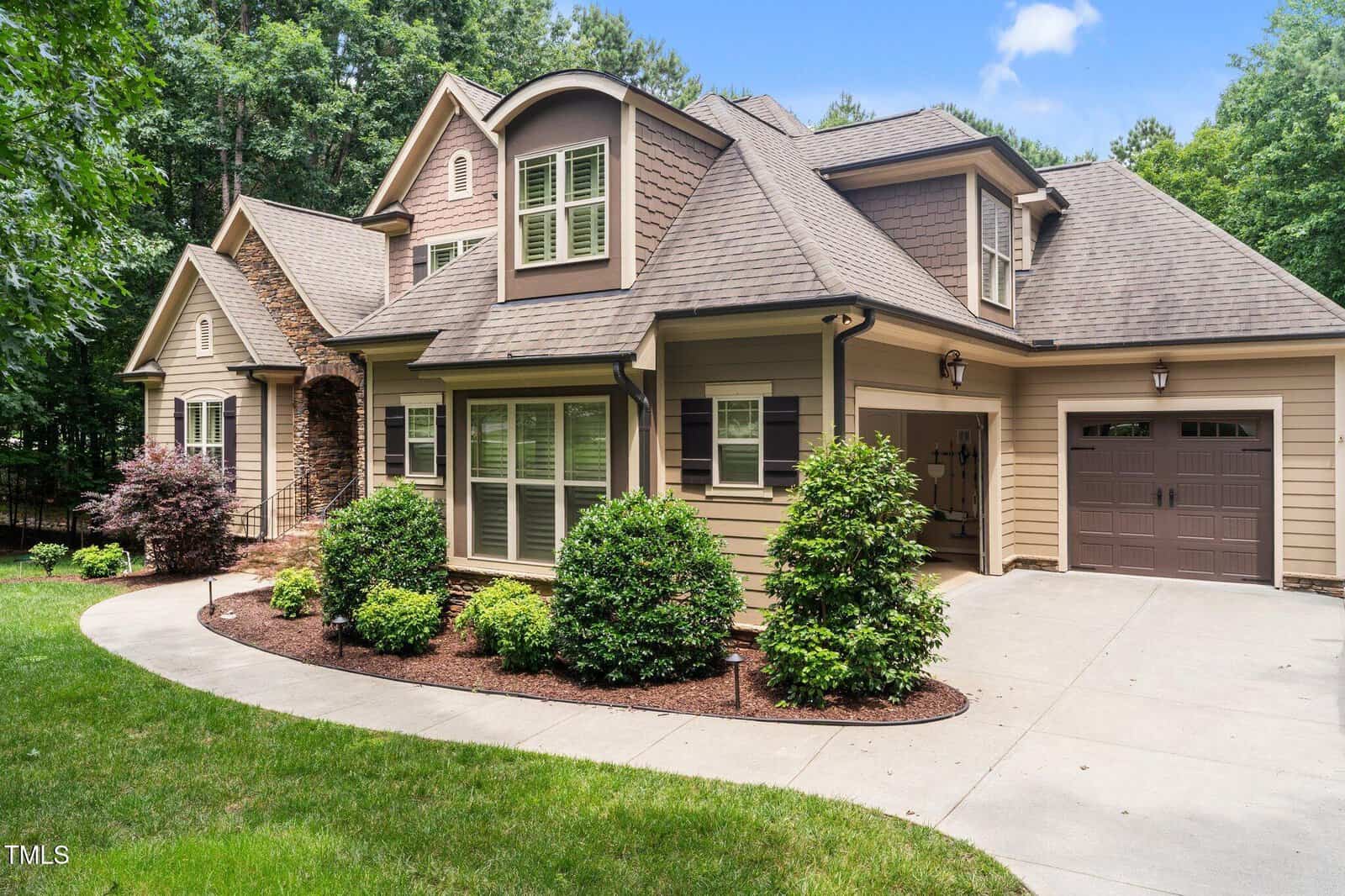 A two-story suburban house with tan siding, multiple gables, large windows, and an attached garage. The house is surrounded by shrubs, grass, and trees, with a driveway curving in front.