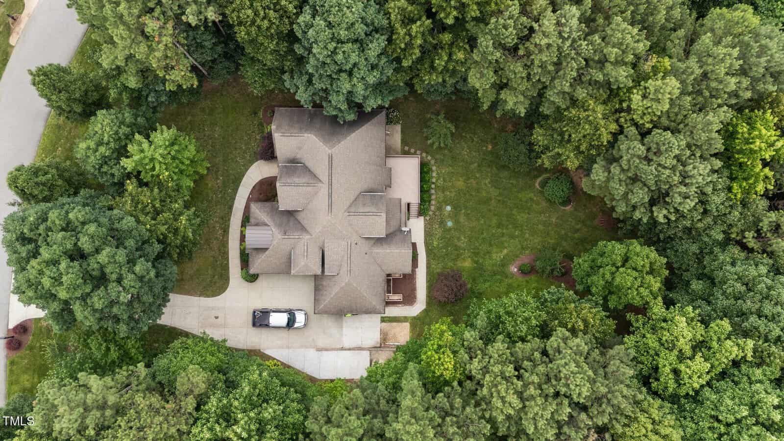 Aerial view of a two-story house with a gray roof, surrounded by dense green trees and a well-maintained lawn; a black and white vehicle is parked in the driveway next to the house.