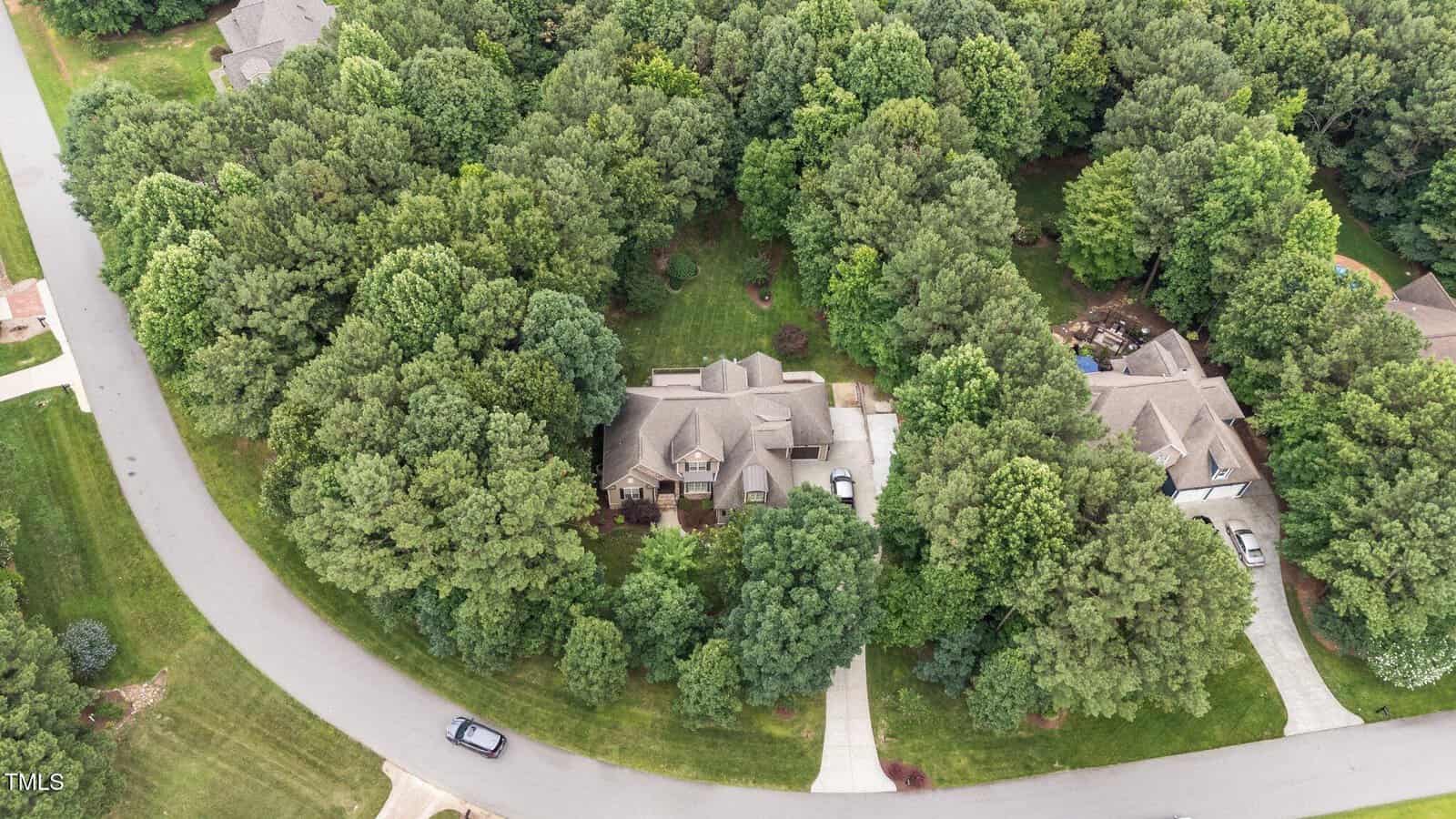 Aerial view of a suburban neighborhood showing two houses with driveways surrounded by dense trees, curved roads, and well-maintained lawns.