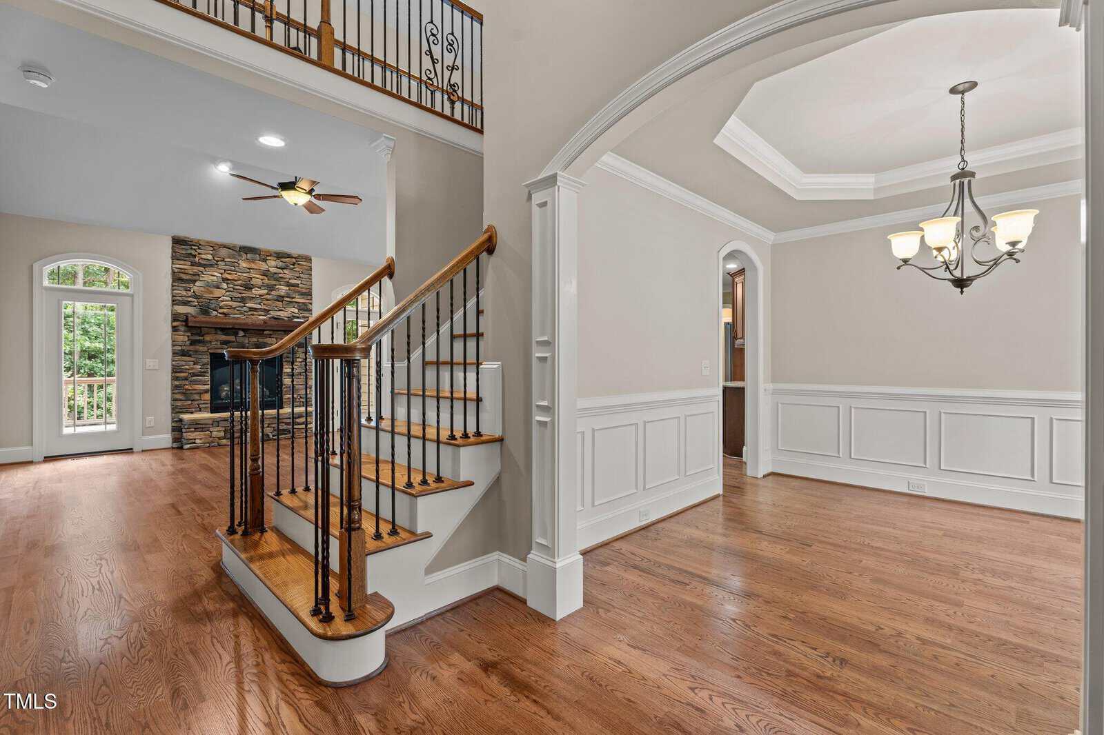 A spacious, unfurnished foyer with wooden floors, a staircase with iron railings, and white wainscoting. An adjacent dining area has a tray ceiling and chandelier. A stone fireplace is visible in the living room.