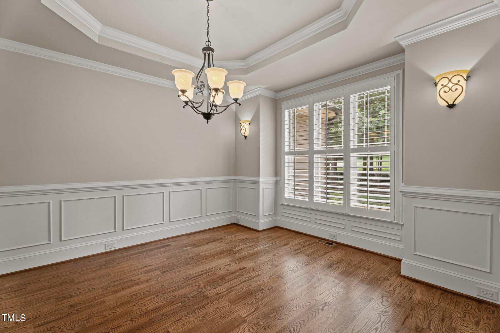 An empty dining room with hardwood floors, beige walls with white wainscoting, a tray ceiling, a chandelier, two wall sconces, and three large windows with white shutters.