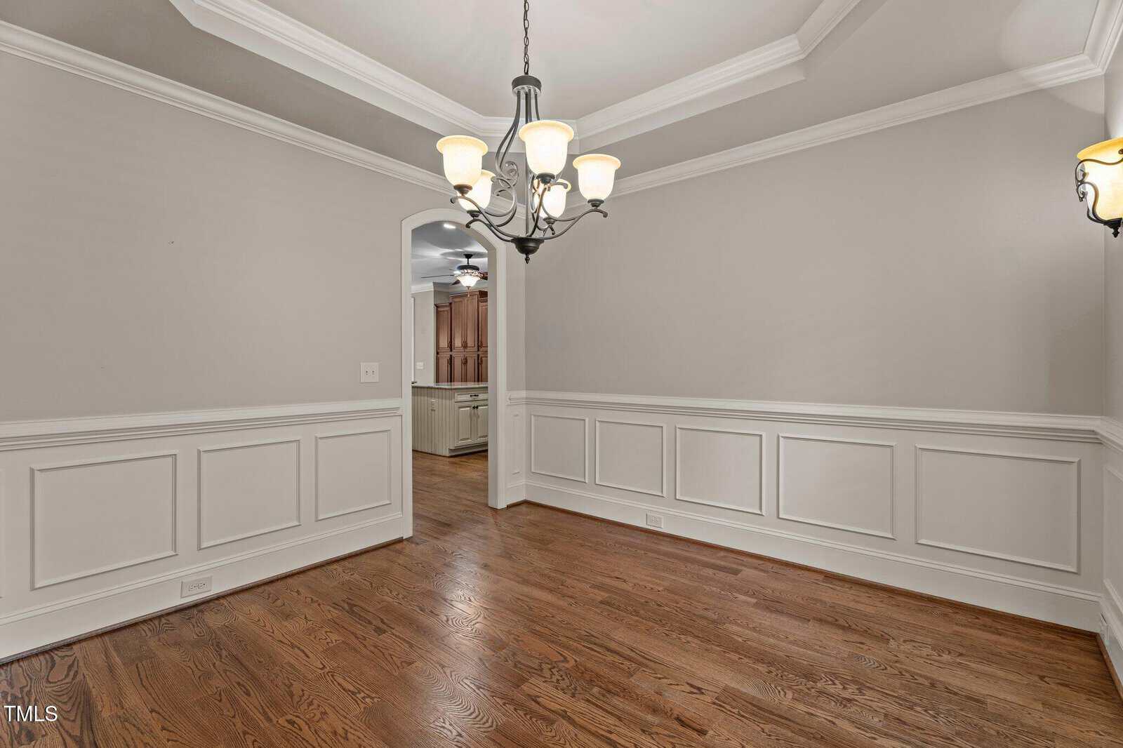 An empty dining room with light beige walls, white wainscoting, hardwood floors, a tray ceiling, and a central chandelier; an open doorway leads to another room with similar decor.