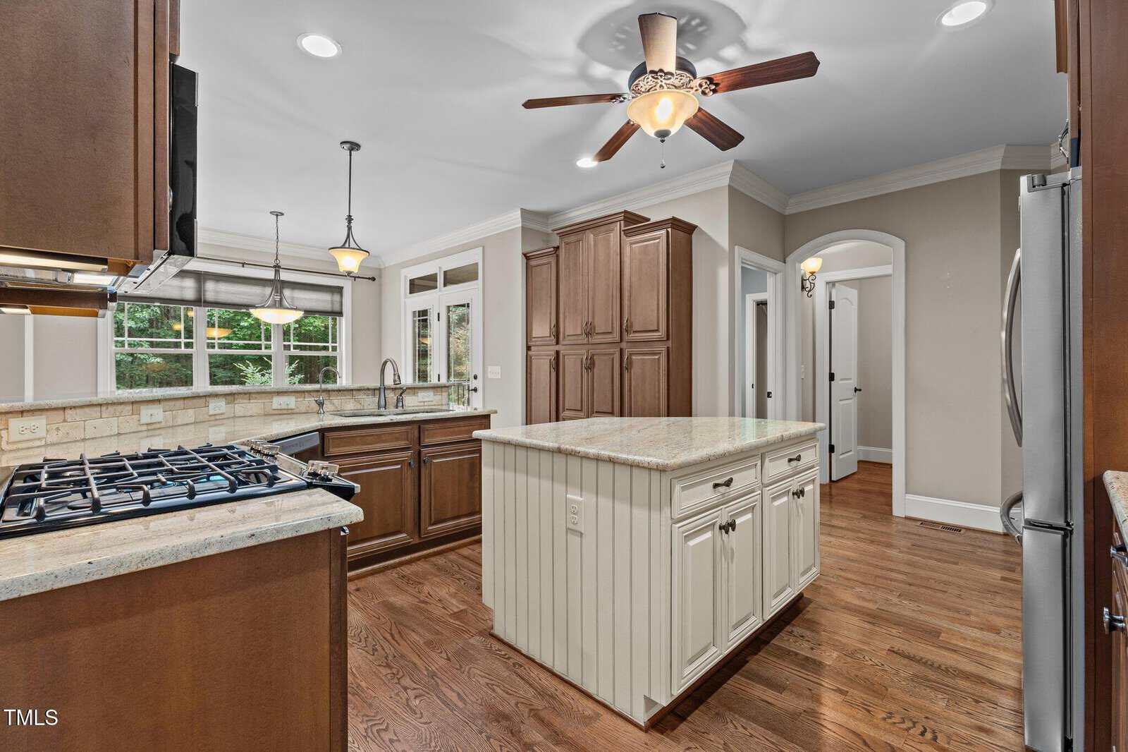 A spacious kitchen with wooden cabinets, a central island with drawers, stainless steel appliances, a ceiling fan with light, and hardwood floors. Large windows provide an outdoor view and natural light.
