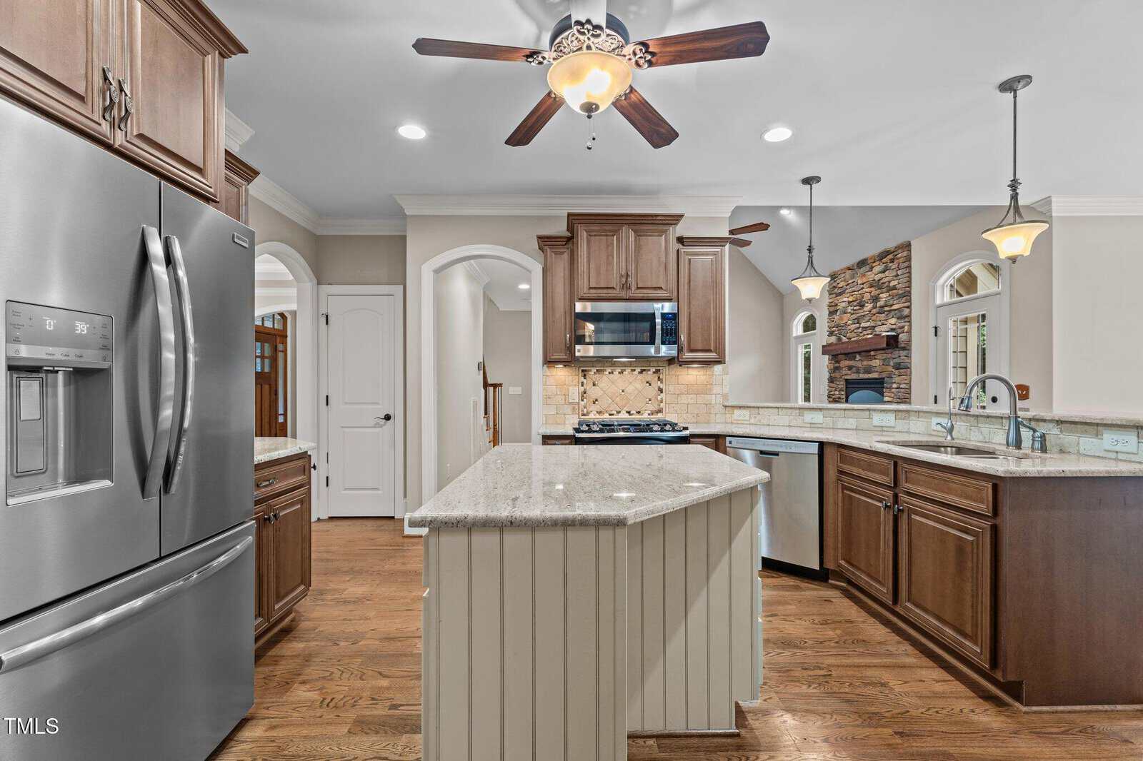 Modern kitchen with stainless steel appliances, granite countertops, wooden cabinets, and a central island. Ceiling fans and pendant lights hang above. An open archway leads to a living room with a stone fireplace.