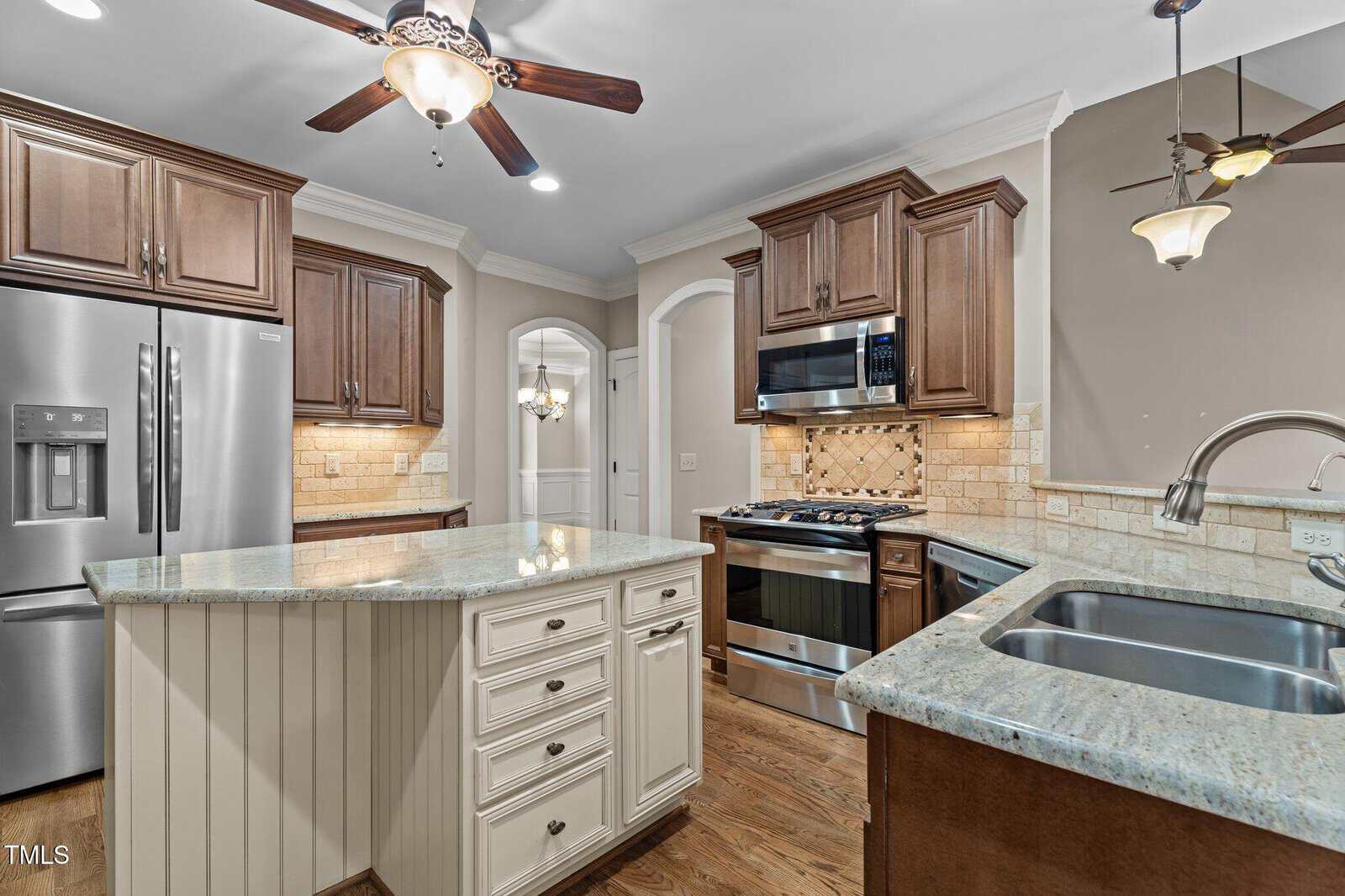 Modern kitchen with brown cabinets, stainless steel appliances, a center island with drawers, granite countertops, double sink, ceiling fans, and pendant lighting. Light-colored backsplash and wooden floors complete the look.