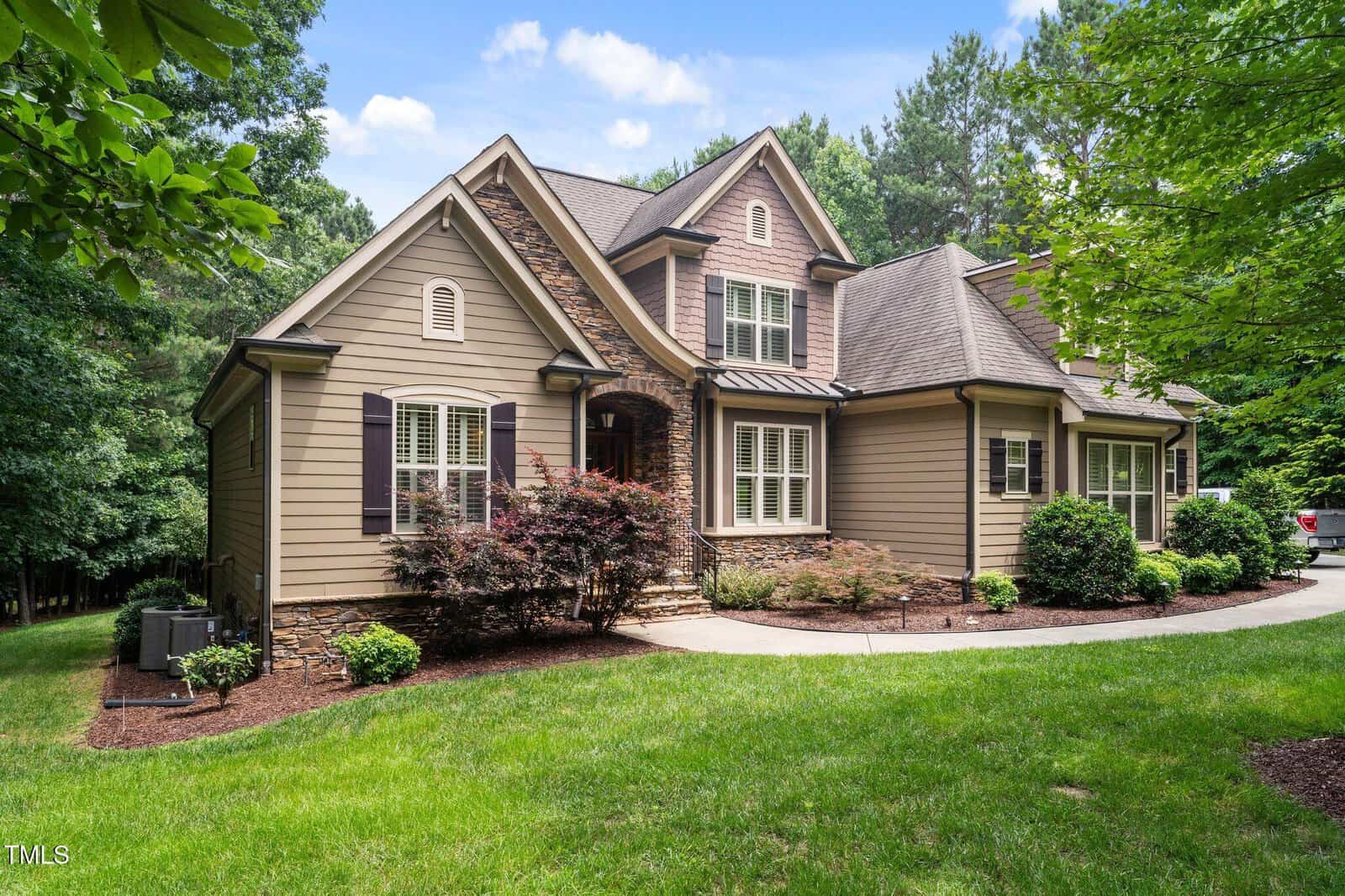 A two-story suburban house with beige siding, dark shutters, stone accents, and a gabled roof sits beside a curved driveway, surrounded by green grass, shrubs, and mature trees on a sunny day.