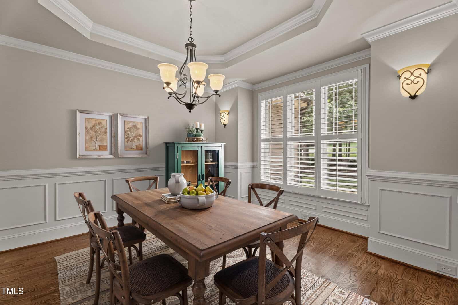 A dining room with a wooden table set for six, wooden chairs, a fruit bowl, a bottle of wine, and glasses. Large windows with white shutters let in daylight; framed art and a green cabinet decorate the room.