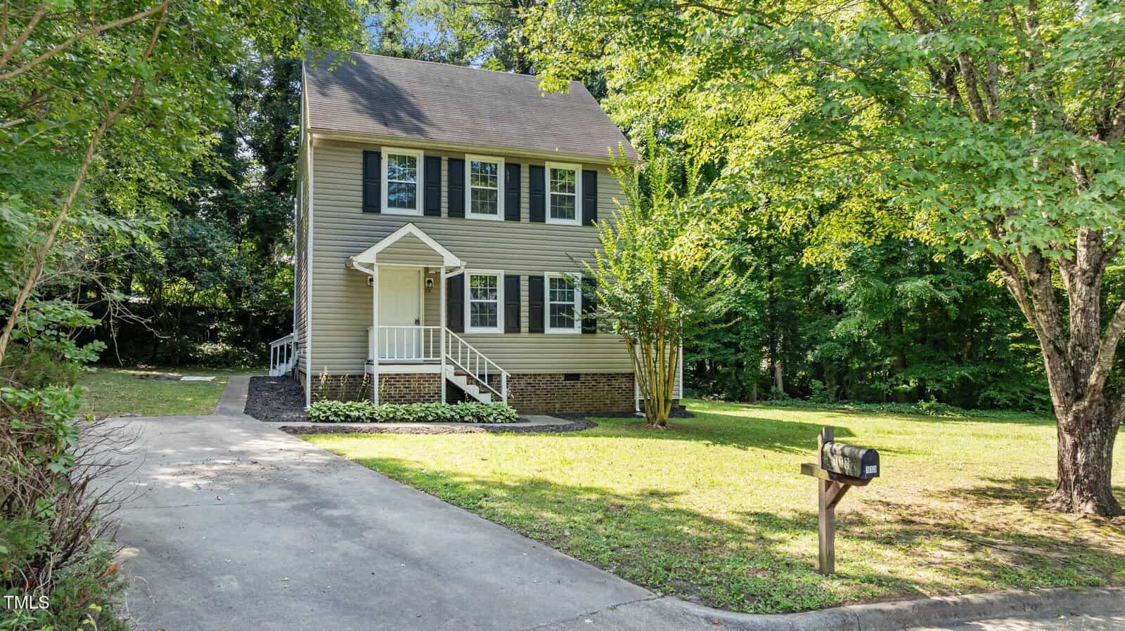 A two-story beige house with black shutters and a covered front porch, surrounded by grass and mature trees. A concrete driveway leads to a mailbox at the edge of the front yard.