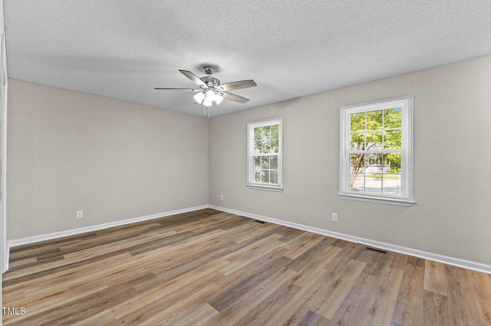 Empty room with light-colored walls, wood-style flooring, a ceiling fan with lights, and two windows letting in natural light. The windows show some greenery outside.