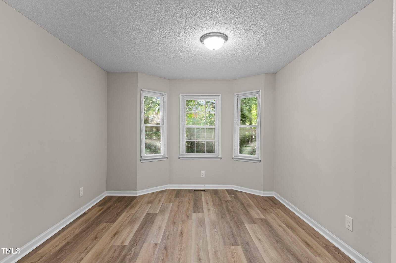 Empty room with light gray walls, wood-look flooring, and a ceiling light. There are three tall windows in a bay window alcove, letting in natural light and showing green trees outside.
