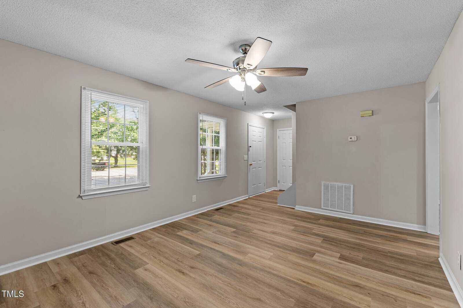 A bright, empty room with light wood flooring, beige walls, two windows, a white front door, a white interior door, a ceiling fan with lights, and a vent cover on the wall.