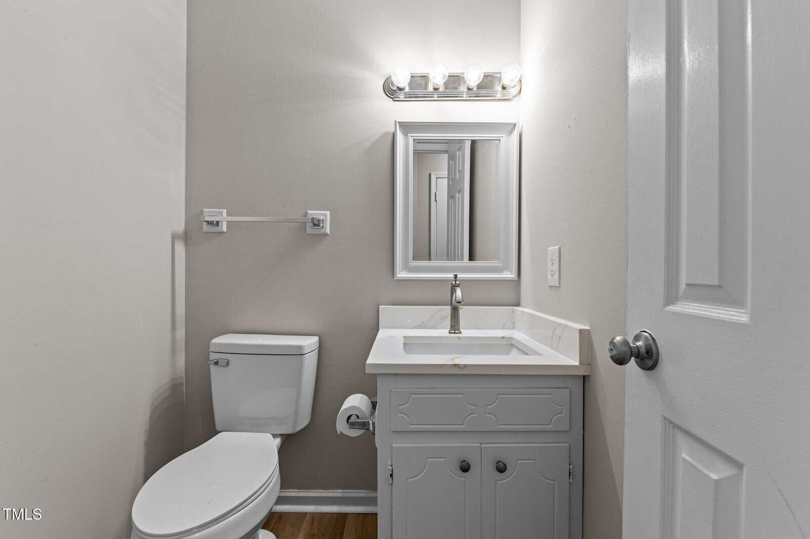 A small bathroom with a white toilet, a white vanity with a sink, a wall-mounted mirror, overhead lighting, and a closed white door. The walls are light-colored and the floor appears to be wood.