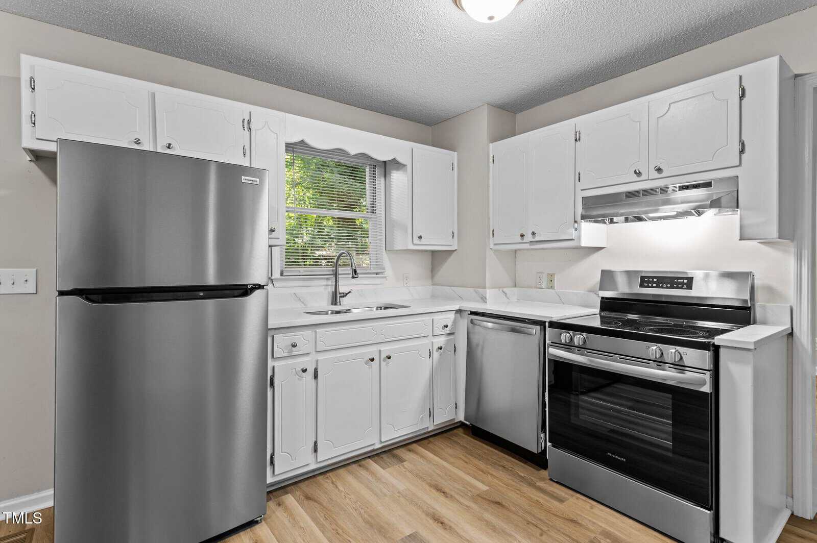 A modern kitchen with white cabinets, stainless steel refrigerator, dishwasher, and oven, a sink under a window, white countertops, and light wood flooring.