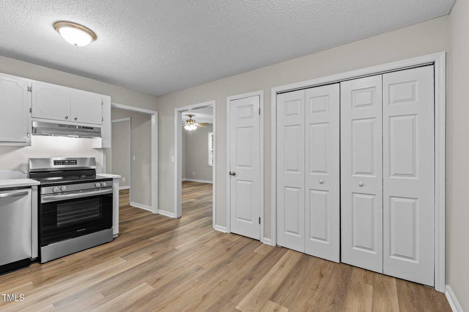 A kitchen with light-colored walls, white cabinets, a stainless steel stove and dishwasher, wood-style flooring, and two sets of white double doors. An open doorway leads to another room with a ceiling fan.