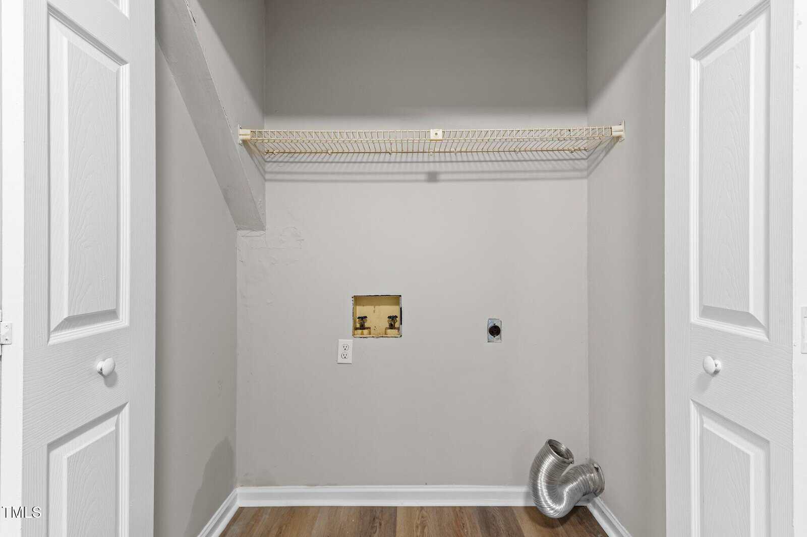 Empty laundry closet with light gray walls, open double doors, a wire shelf, hookups for a washer and dryer, exposed vent hose on the floor, and wood-style flooring.