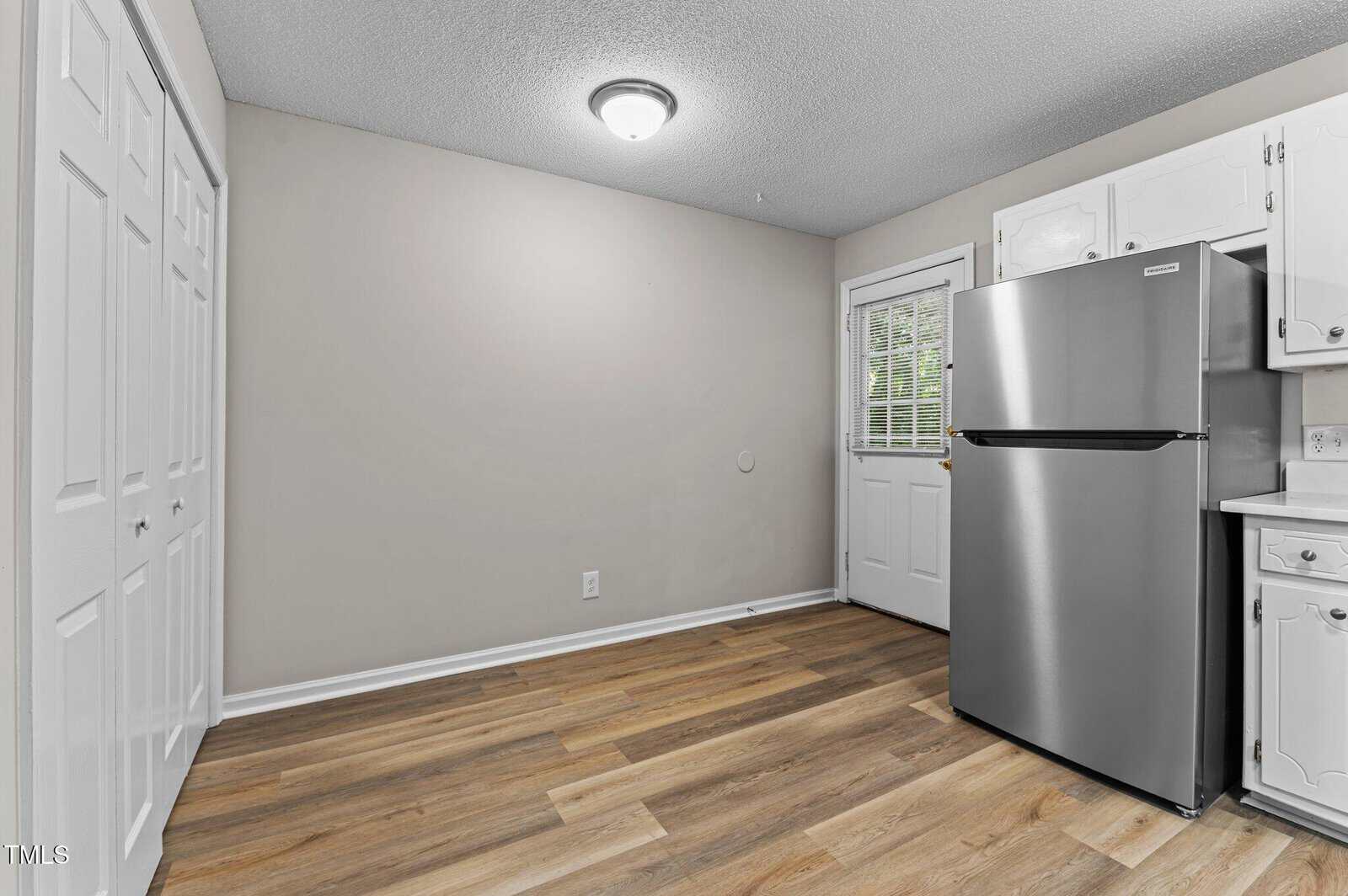 A kitchen with light wood flooring, white walls, white cabinets, a stainless steel refrigerator, and a door with a window leading outside. A ceiling light fixture is centered on the textured ceiling.