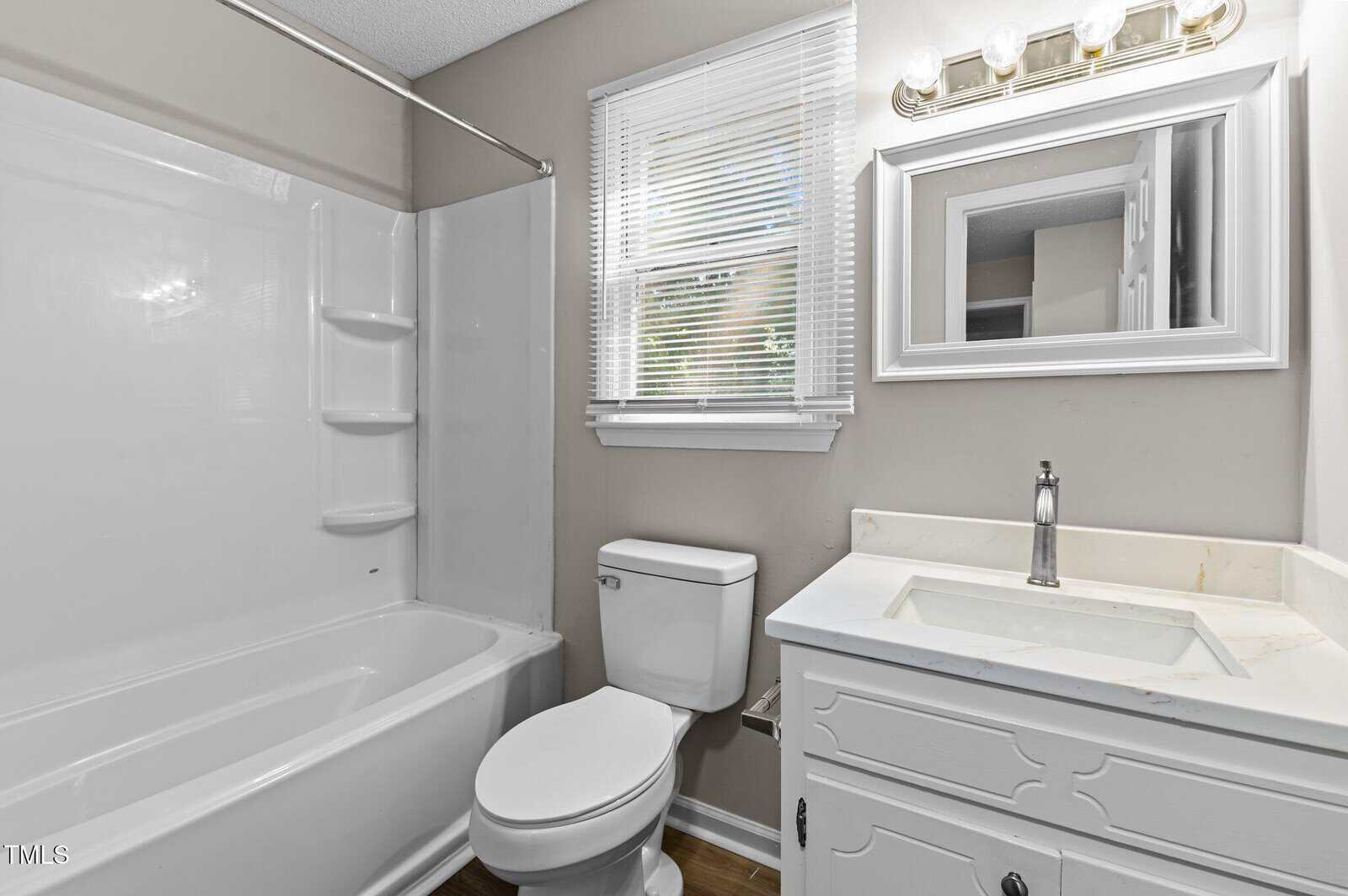A clean bathroom with a white bathtub-shower combo, toilet, and white vanity with a marble countertop and mirror above. A window with blinds lets in natural light.