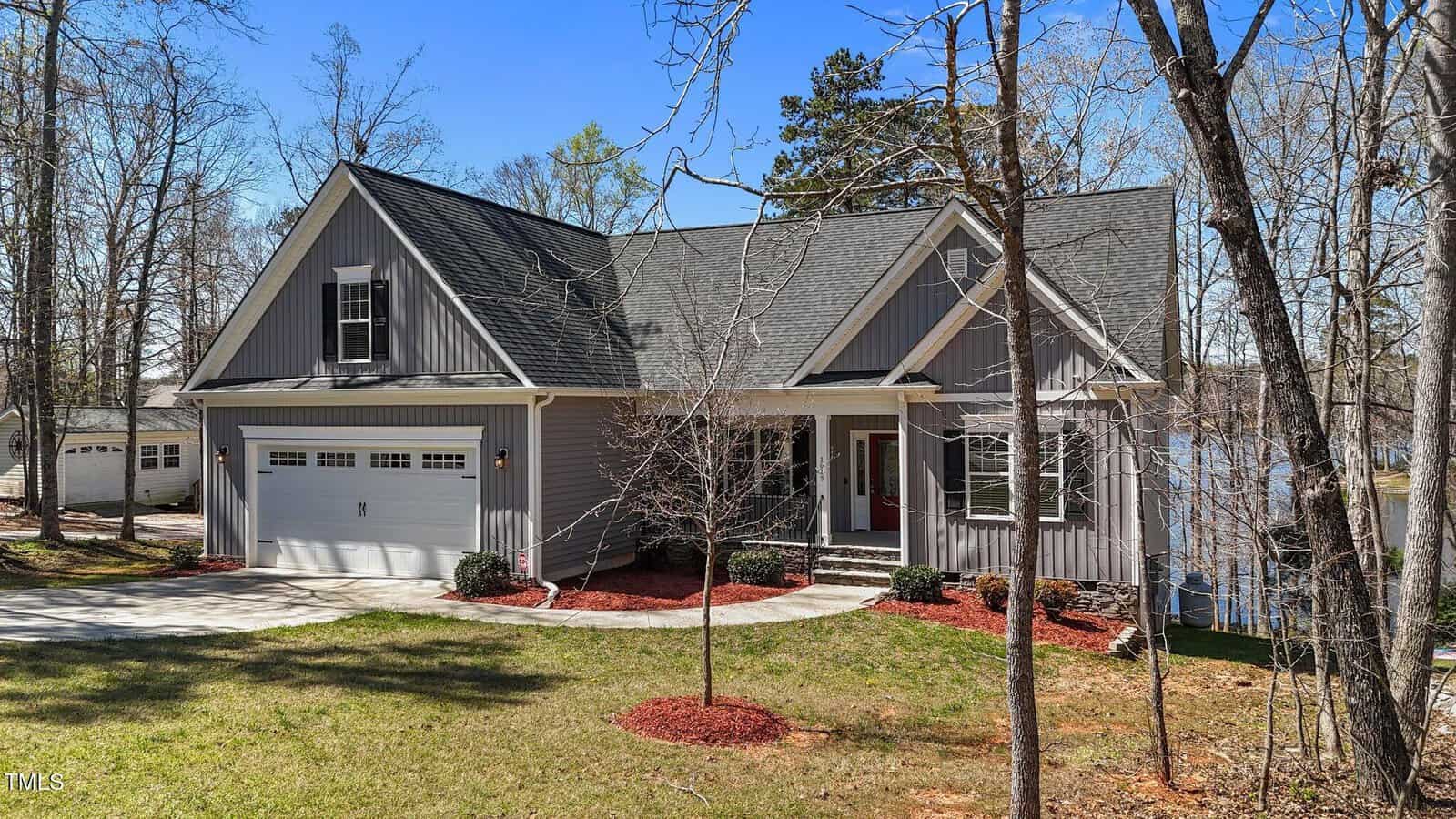 A gray, two-story house at 1013 Sagamore Drive, Louisburg, with a front porch, attached two-car garage, and gabled roof is nestled among trees and a grassy yard, with a lake visible in the background.