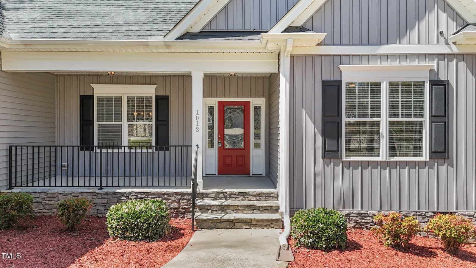 A house exterior with gray siding, white trim, a red front door, stone steps, black shutters, a covered porch with railing, and neatly trimmed bushes along a mulched walkway.