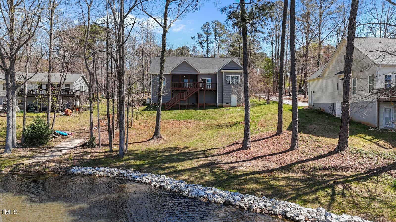 A gray house with a wooden porch and steps sits on a grassy slope by a small lake, surrounded by trees. Neighboring houses are visible on both sides under a clear blue sky.