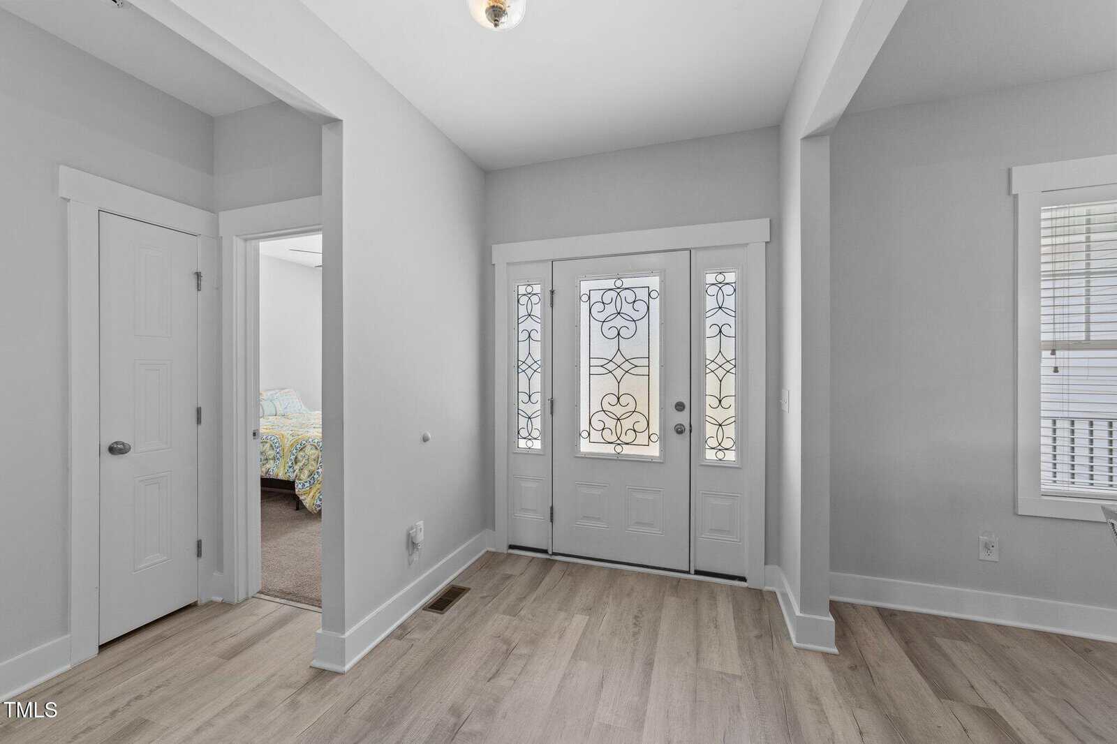 Bright entryway with light wood floors, white walls, and a front door featuring decorative glass panels. An open doorway reveals a bedroom with a bed, and a window with blinds is visible to the right.