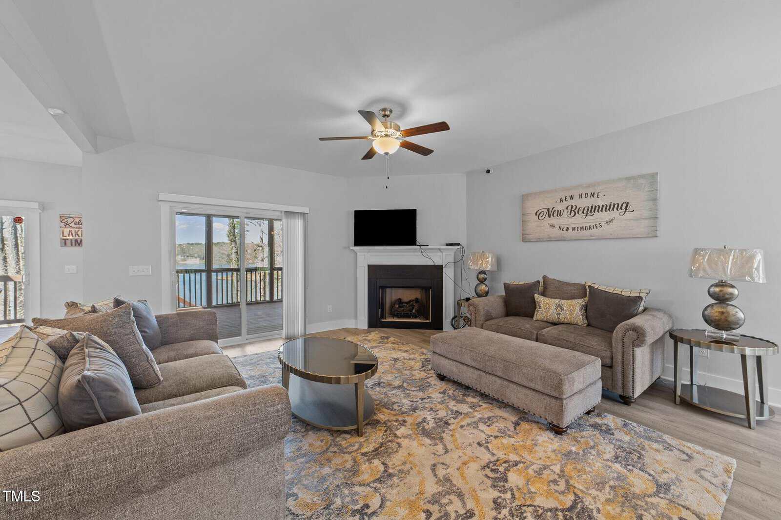 A modern living room with gray sofas, a patterned rug, glass coffee table, fireplace, and TV. Sliding doors open to a balcony with a lake view, and a wall sign reads, "New Home, New Beginning.
