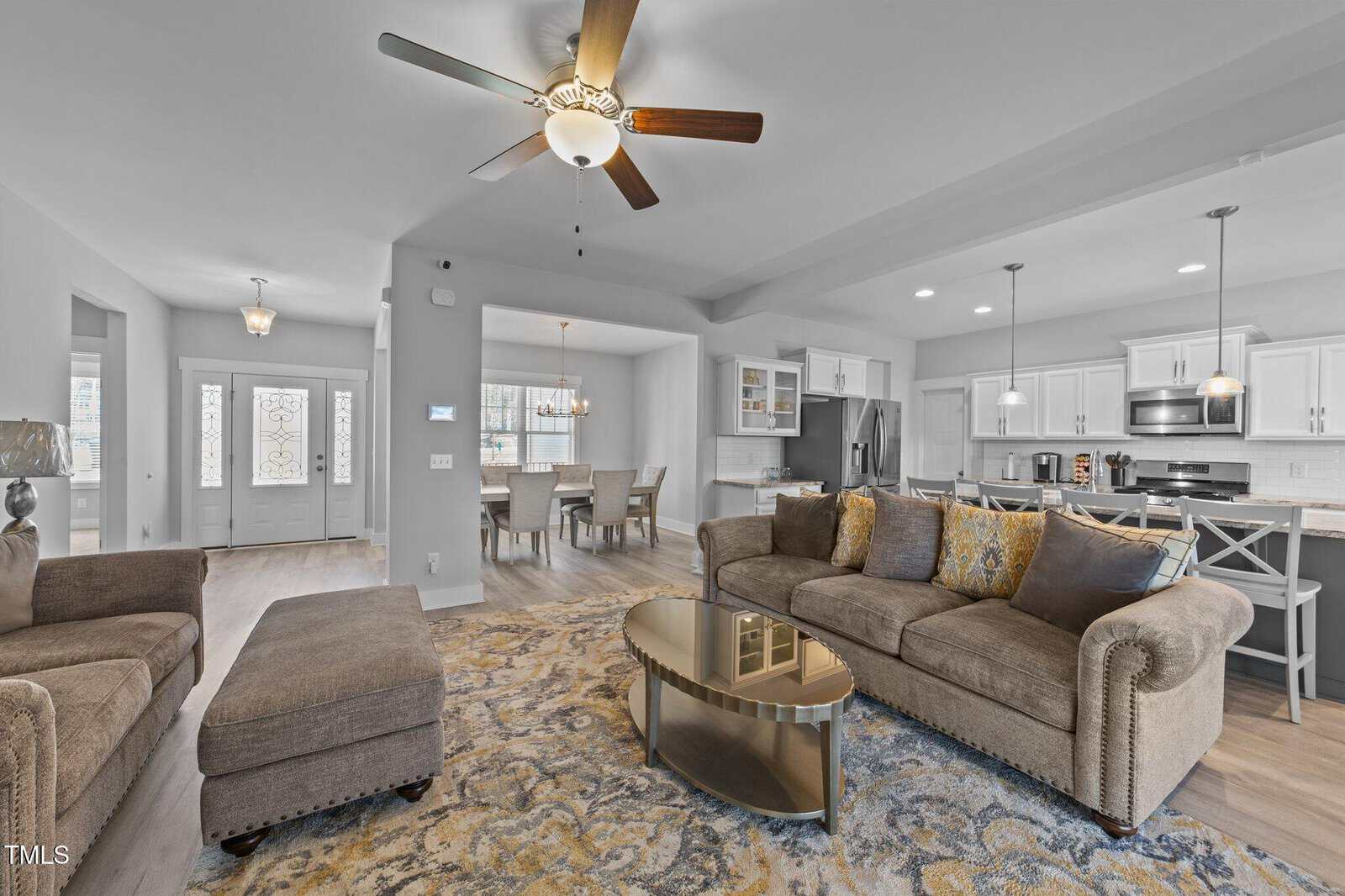 Modern open-concept living room with neutral tones, plush seating, a patterned rug, and a round glass coffee table. The space connects to a kitchen with stainless steel appliances and a dining area. Ceiling fans and natural light brighten the room.