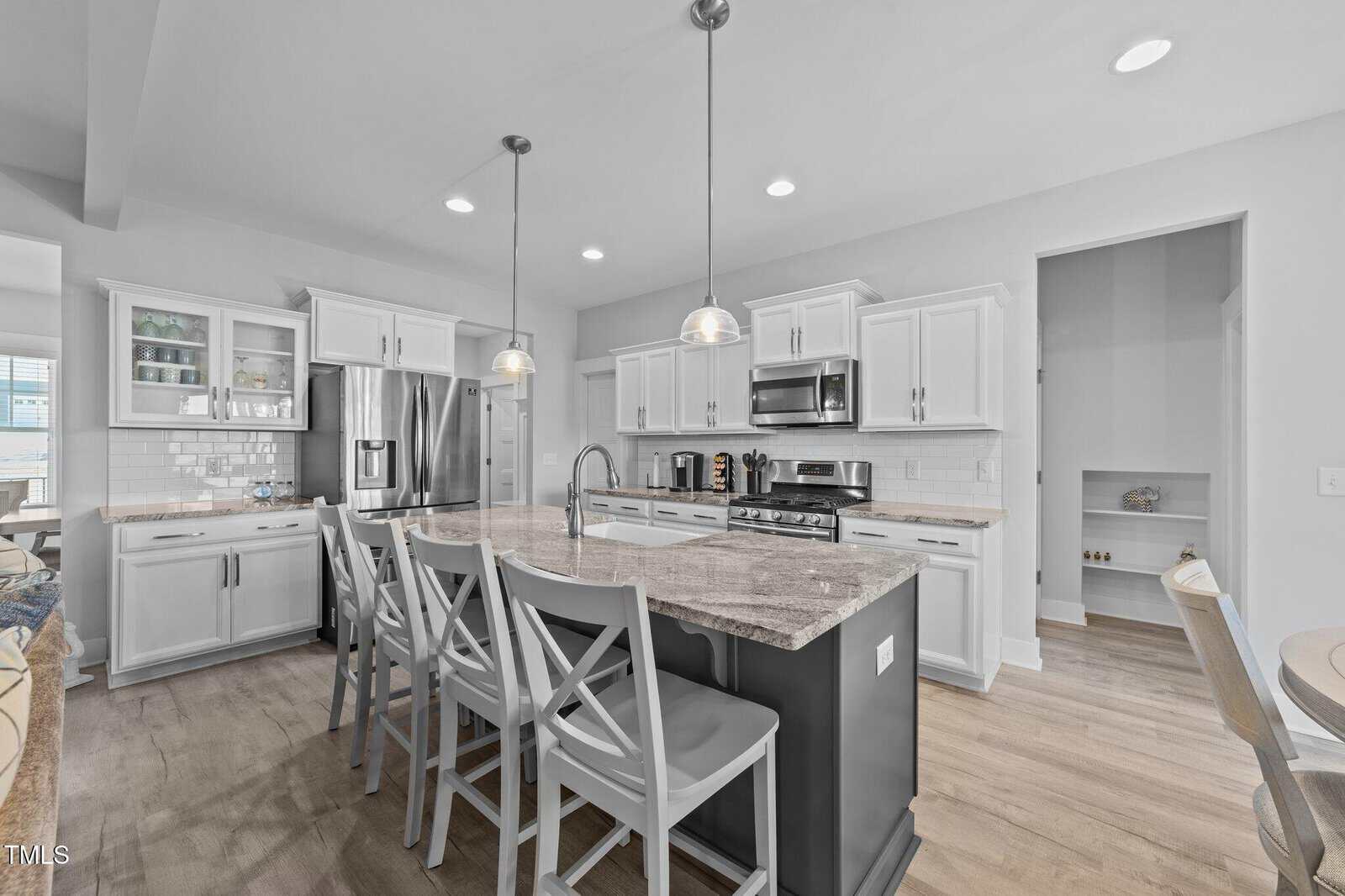 Modern kitchen with white cabinets, stainless steel appliances, a large center island with granite countertop, four bar stools, hanging pendant lights, and light wood flooring in a bright, open space.