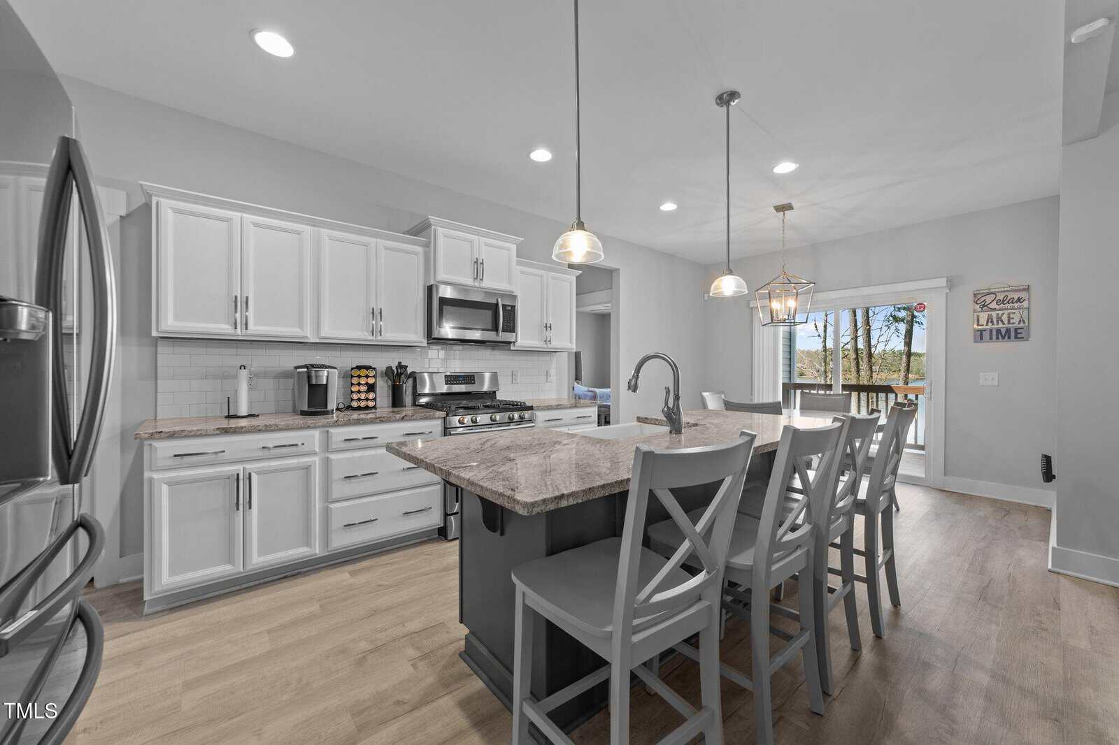 Modern kitchen with white cabinets, stainless steel appliances, a large island with four gray chairs, pendant lighting, and wood floors. Sliding glass doors lead to a deck with trees visible outside.