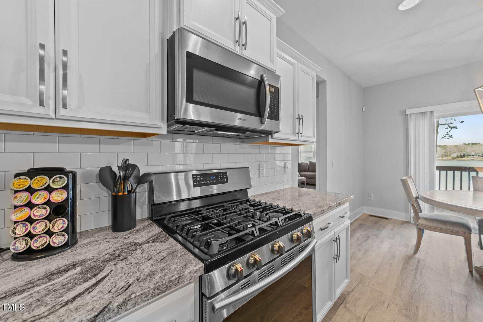 Modern kitchen with stainless steel gas stove and microwave, white cabinets, marble countertop, knife set, and coffee pod holder; dining area and large window with water view in background.