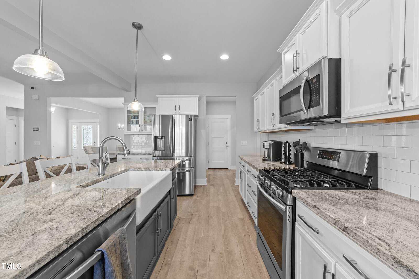 Modern kitchen with stainless steel appliances, granite countertops, white cabinets, a farmhouse sink, light wood flooring, and pendant lights. Dining table with chairs is visible in the background.