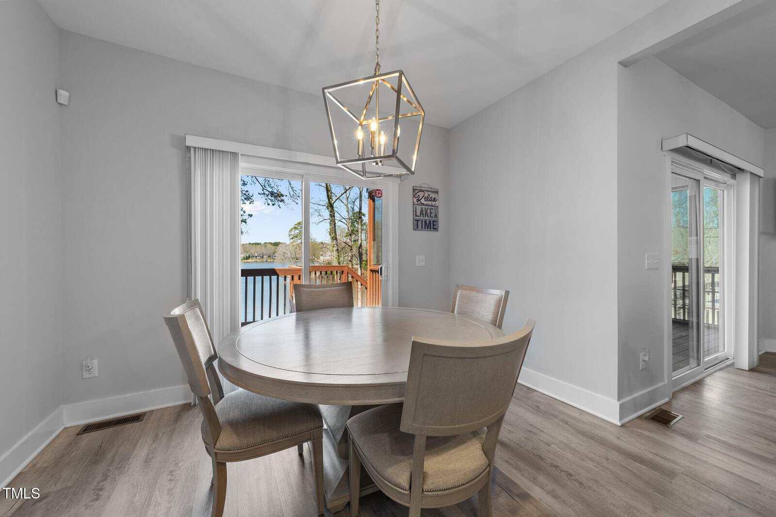Bright dining area with a round wooden table and four chairs, modern chandelier above, large sliding glass doors opening to a deck with a lake view, and light wood floors throughout.