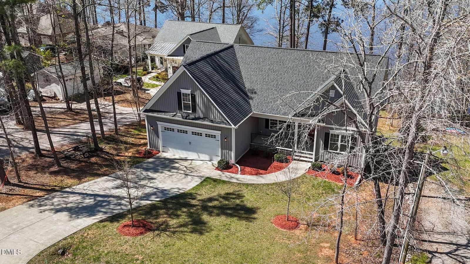A gray two-story house with a front porch, attached two-car garage, and curved driveway sits among tall trees near a lake, with neighboring homes visible and landscaped red mulch beds in the yard.