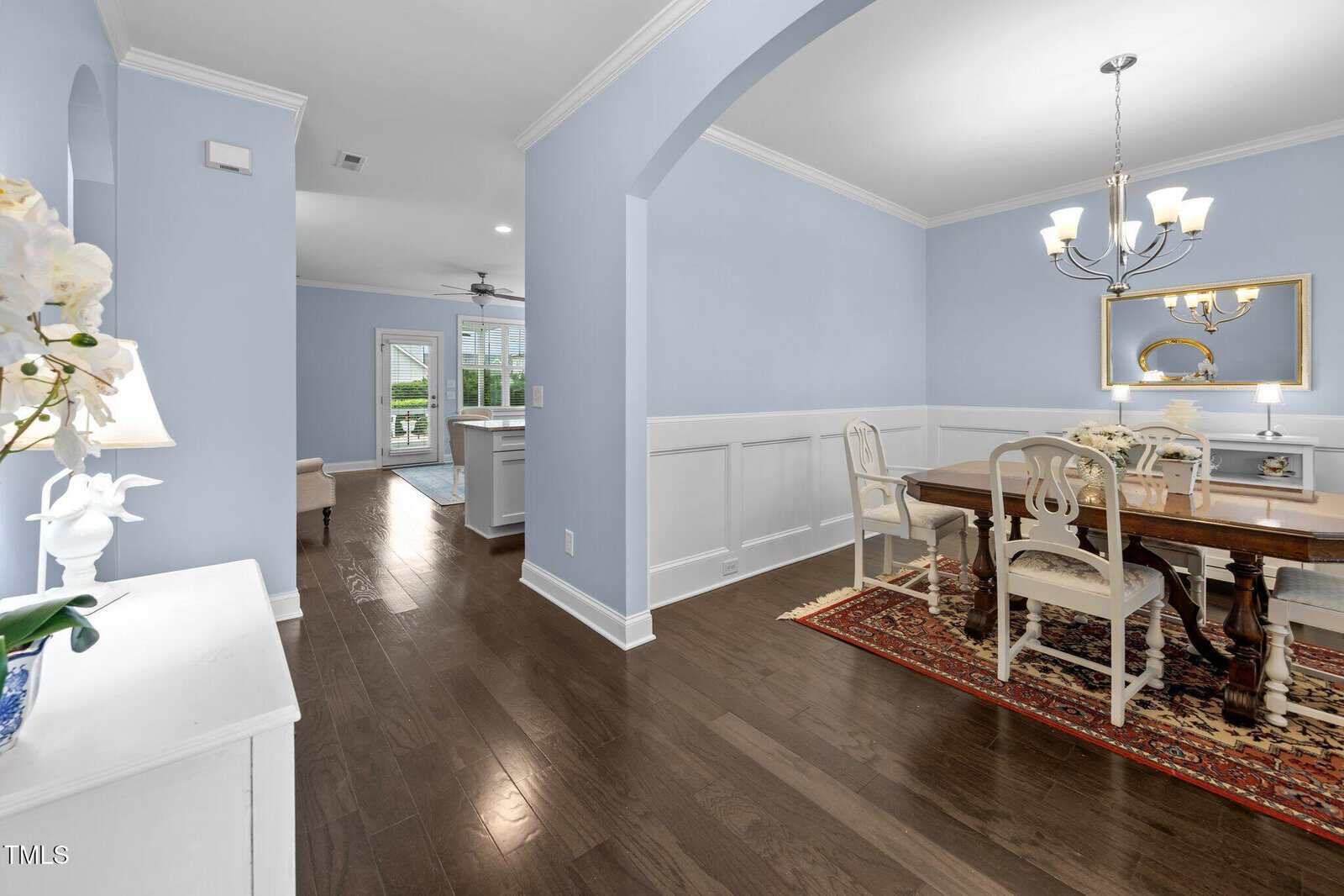 A dining room with a wooden table and chairs, chandelier, decorative rug, and mirror. The space features light blue walls, white wainscoting, and dark hardwood floors, leading into a bright living area and kitchen.
