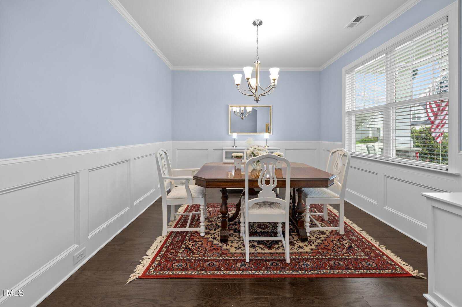 A bright dining room with light blue walls, white wainscoting, a wooden dining table, six white chairs, a red patterned rug, a chandelier, a mirror, and a large window letting in natural light.