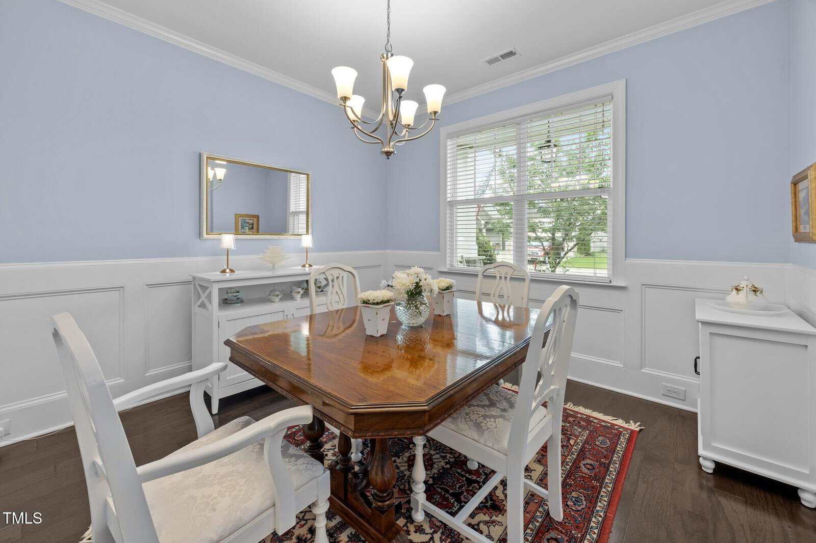 A dining room with pale blue walls, a wood table, white chairs, a chandelier, a floral centerpiece, a sideboard with a mirror, hardwood floors, a patterned rug, and a window with white blinds showing greenery outside.
