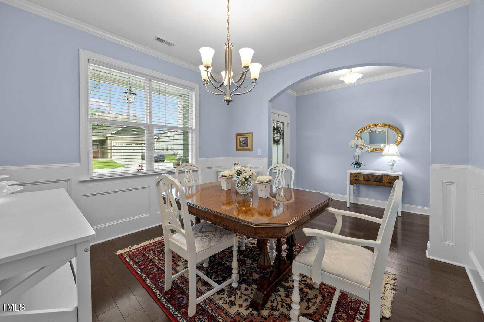 A dining room with light blue walls, white trim, a wooden table with six chairs, a chandelier, a window with blinds, a red-patterned rug, and a sideboard. An archway leads to a hallway with a console table and mirror.