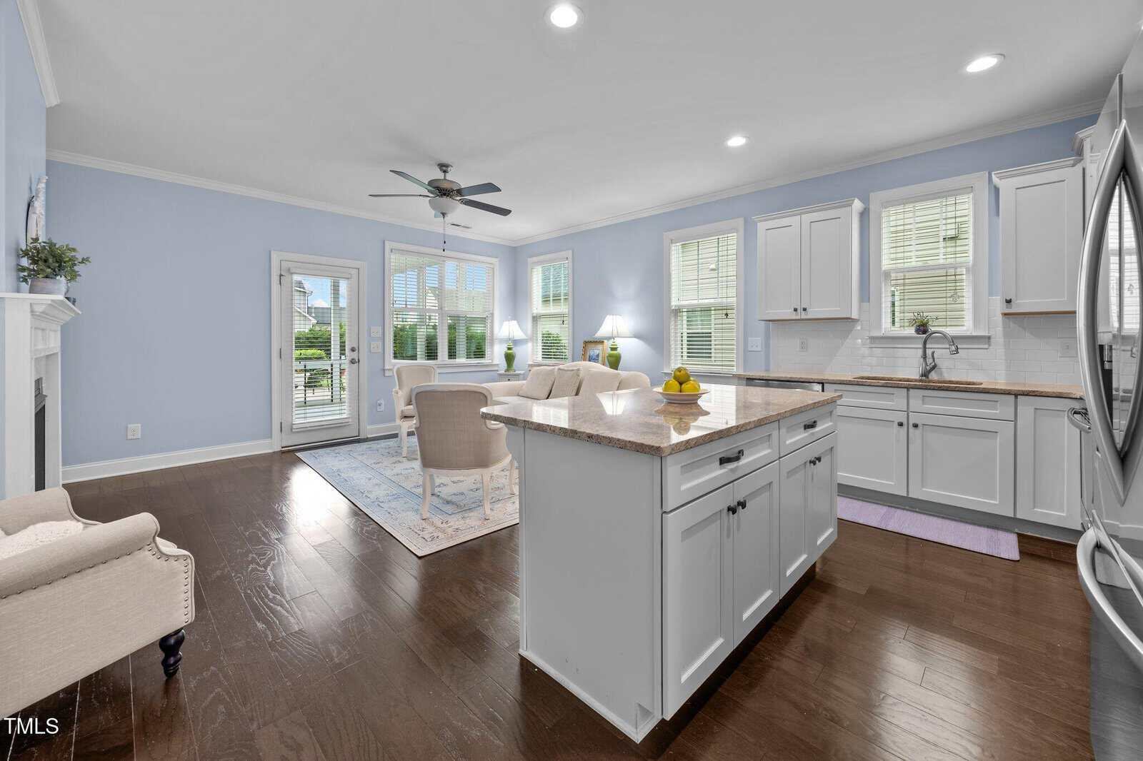 Bright kitchen and living area with pale blue walls, white cabinets, a central island, dark wood floors, and large windows letting in natural light. A ceiling fan and patio door are visible in the background.