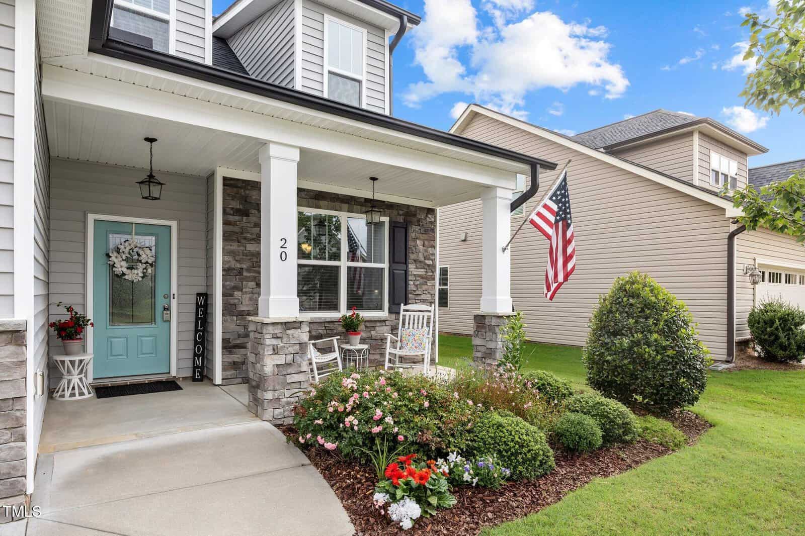A modern suburban house with stone accents, a front porch with white rocking chairs, potted flowers, a welcome sign, and an American flag. A manicured lawn and flower beds surround the entrance.