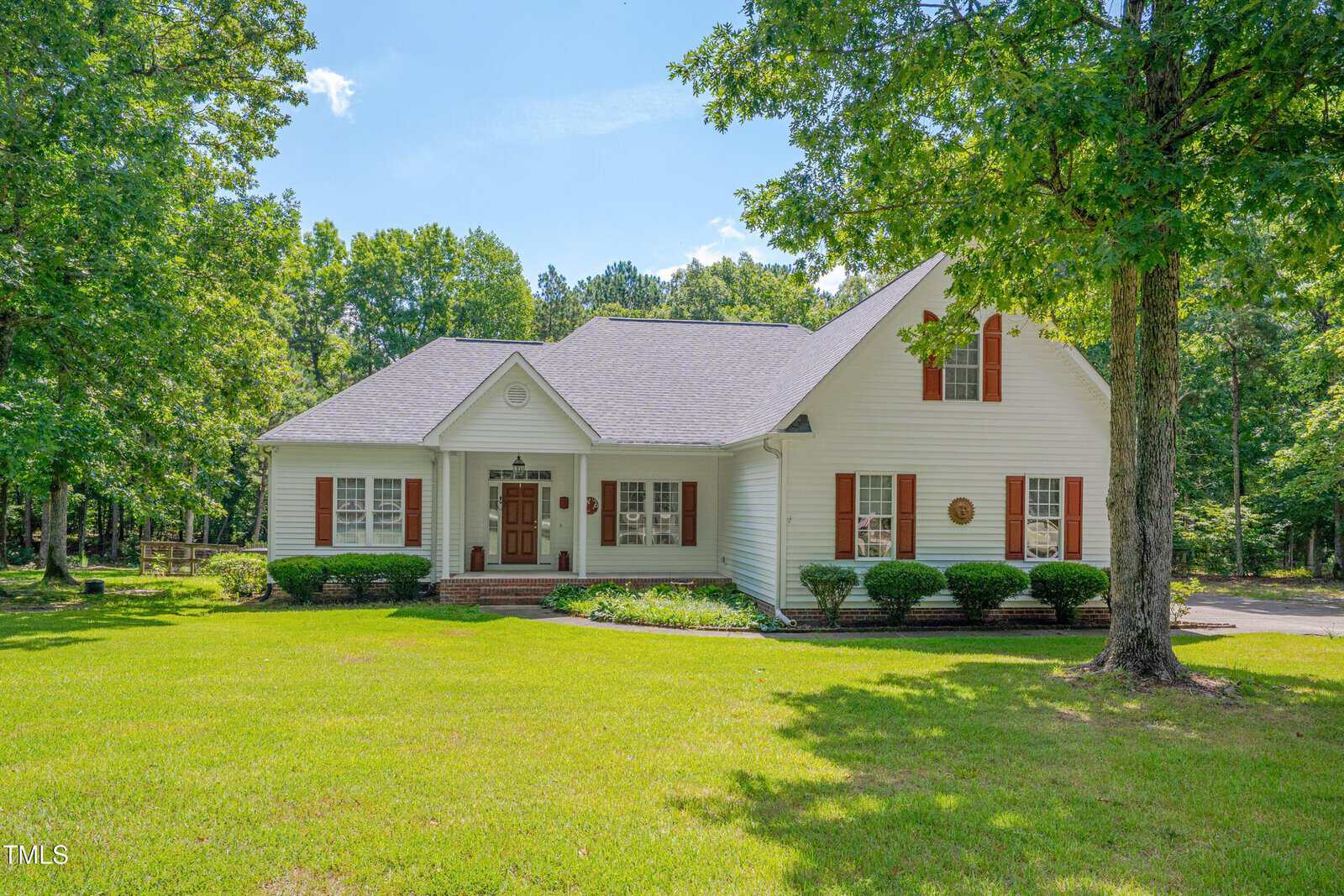 A white single-story house with red shutters, a gable roof, and a large front lawn, surrounded by green trees on a sunny day. Brick steps lead to the front door, and shrubs line the front porch.
