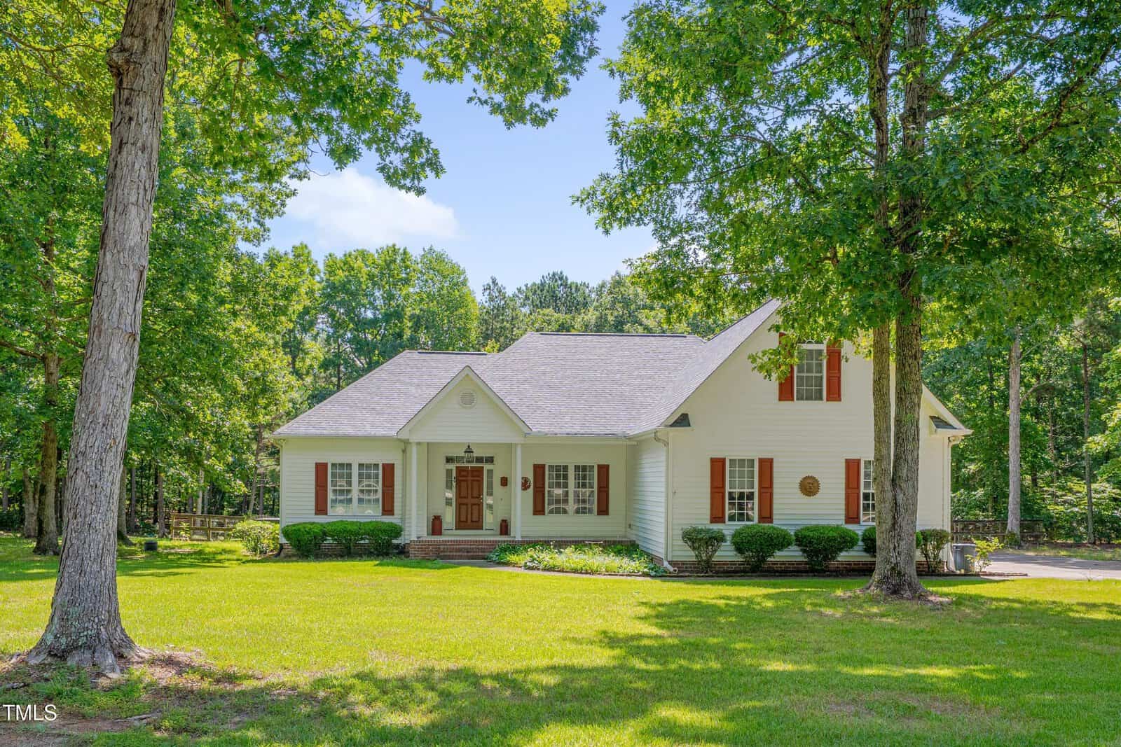 A white two-story house with red shutters, a covered front porch, and manicured shrubs sits amid a spacious, sunny lawn surrounded by tall green trees.