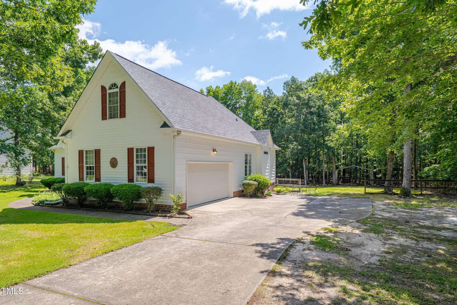A white, two-story house with brown shutters and a gray roof, featuring a double garage and a curved driveway, surrounded by green lawn, shrubs, and tall trees under a blue sky.