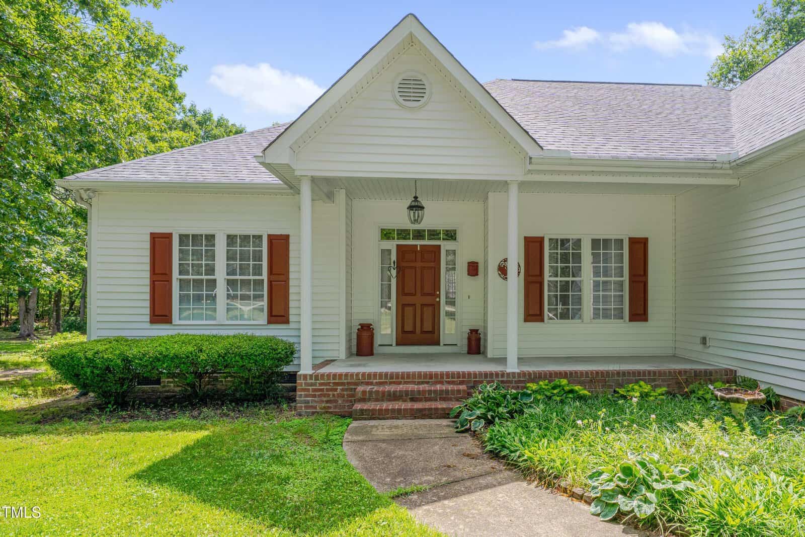 Single-story white house with a covered front porch, brick steps, a wooden front door, red shutters on the windows, and landscaped green lawn and bushes under a partly cloudy sky.