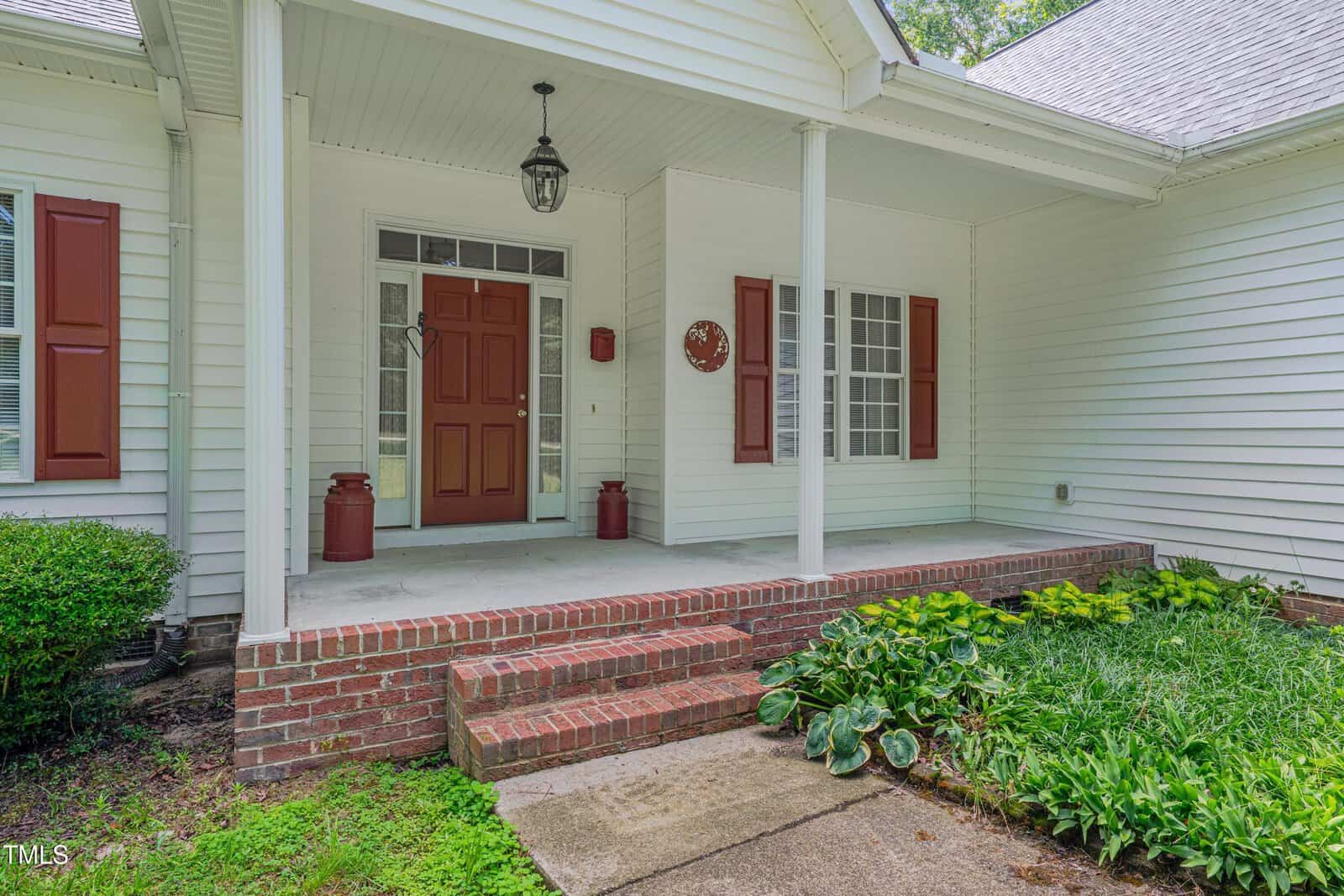 A white house with red shutters and a covered front porch, featuring brick steps, a red front door, two vintage milk cans, and green plants by the walkway.