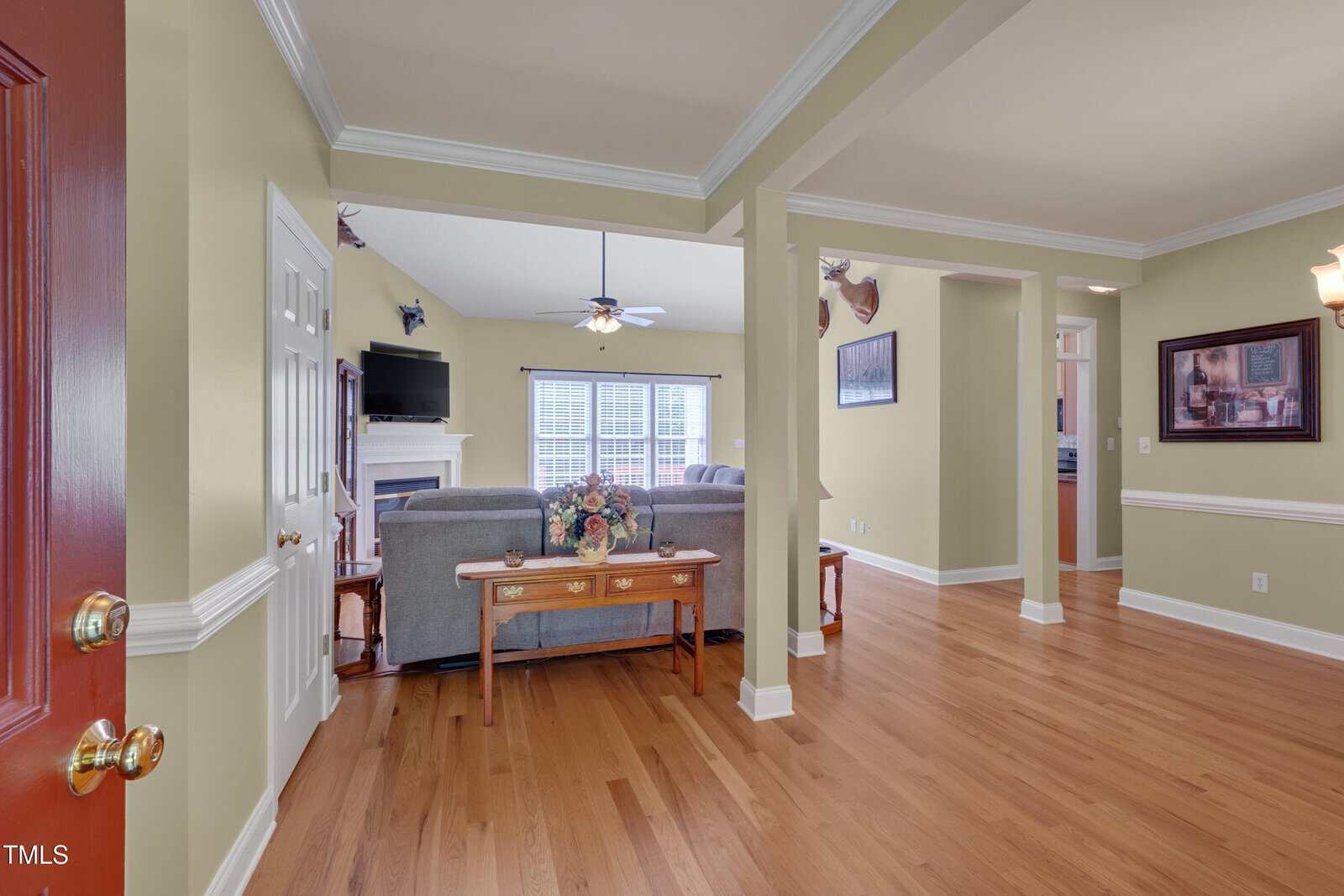 Entryway view into a living room with hardwood floors, yellow walls, and white trim. A sofa and table are centered, fireplace and TV in the background, ceiling fan, windows, and wall art visible.