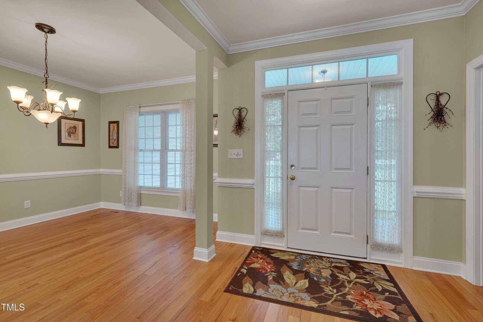 A front entryway with a white door, sidelights, a transom window, a floral doormat, hardwood floors, light green walls, and an adjacent dining area with a window, chandelier, and framed artwork.