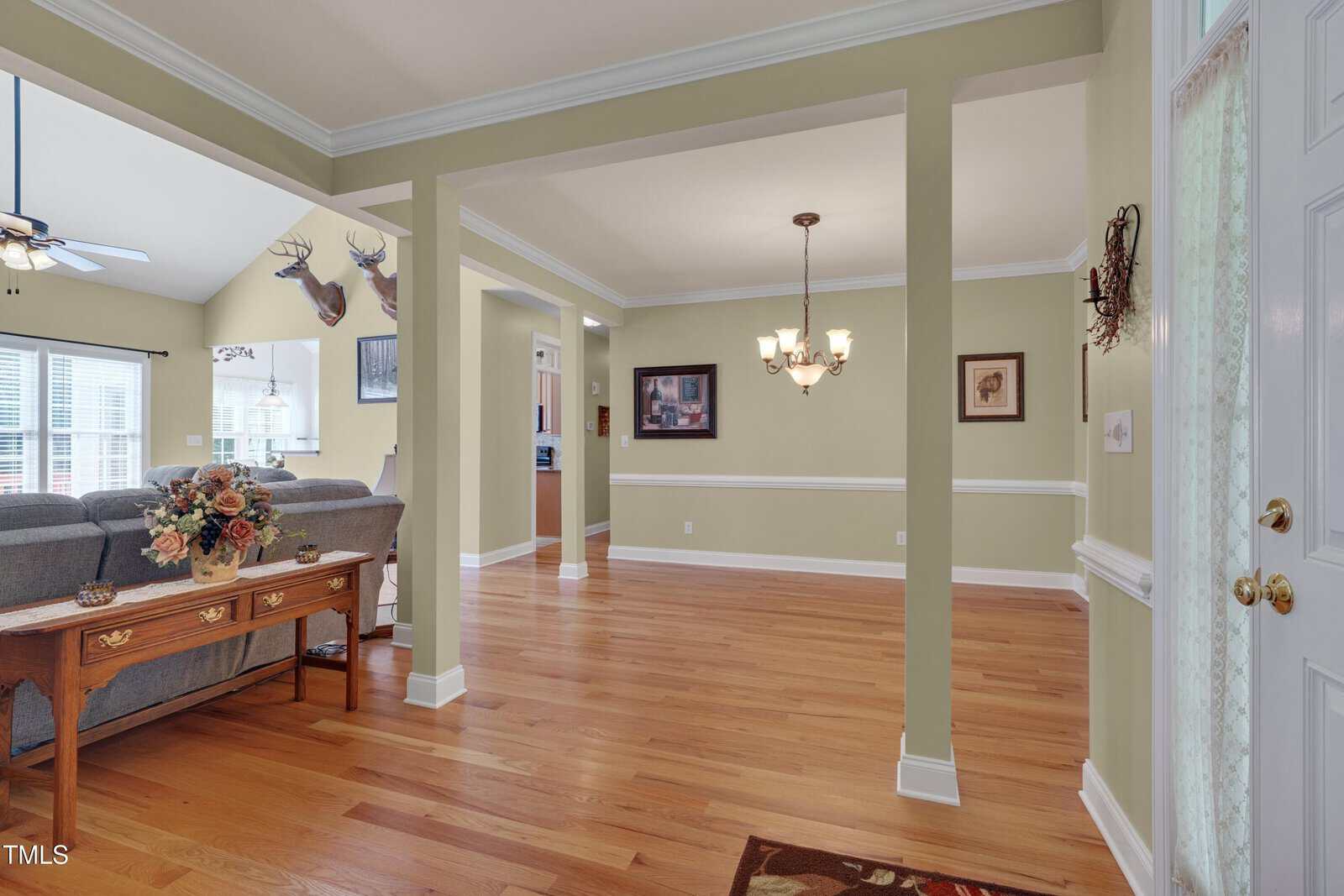 A living room with hardwood floors, beige walls, and white trim. There is a chandelier, a sofa near a window, mounted deer heads, and framed art. A wooden table with a flower arrangement stands by the entryway.