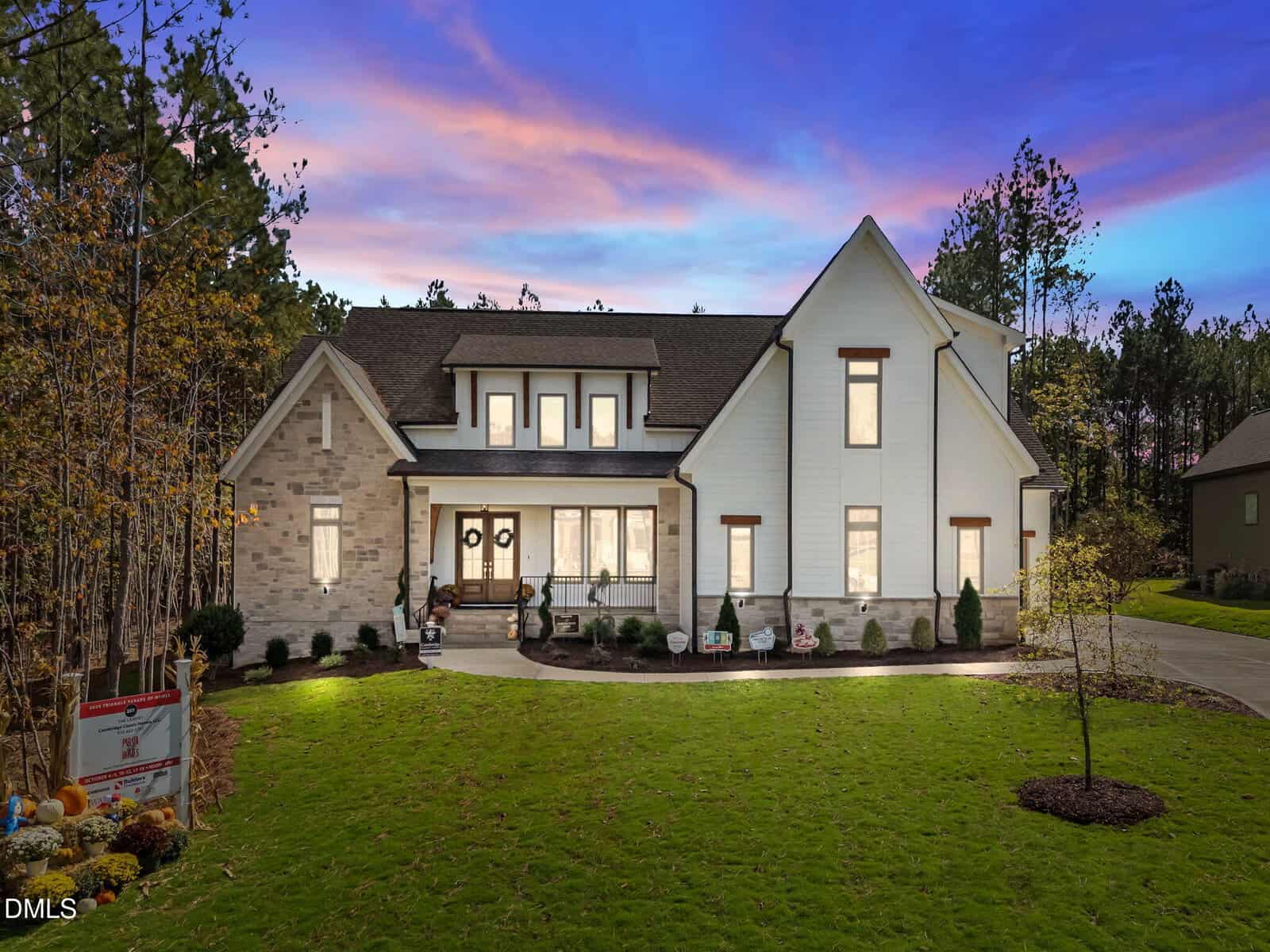 A large two-story house with white siding, brick accents, and multiple lit windows sits on a neatly landscaped lawn at dusk in Youngsville, with trees and a colorful sky in the background. For sale signs are visible near the front yard of this beautiful home at 70 Spanish Oak Drive.