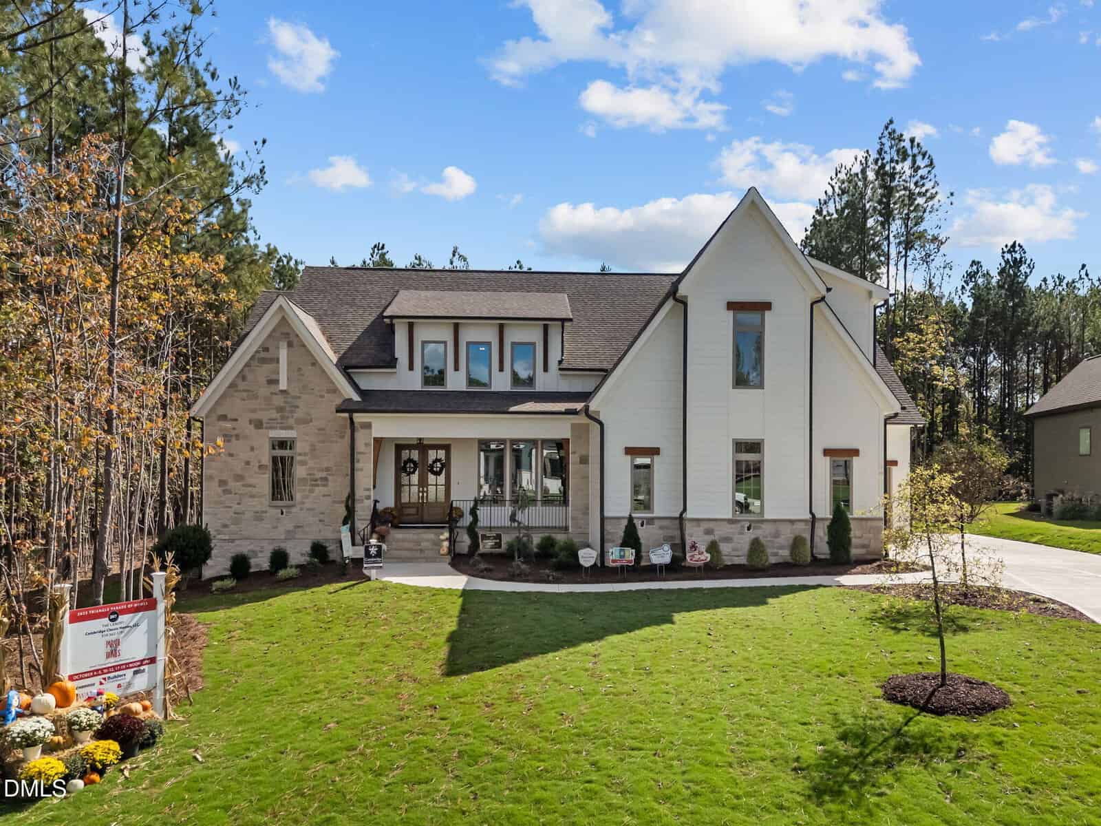 Two-story modern suburban house with brick and white siding exterior, a covered front porch, and a well-kept lawn. A driveway curves to the right, and trees surround the property. Decor and a sign are near the front walkway.