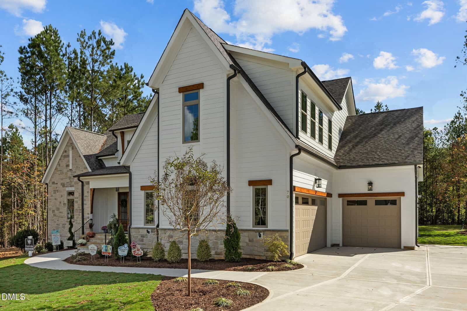 A modern two-story house with white siding, stone accents, a front porch, three-car garage, landscaped yard, and trees in the background under a blue sky with clouds.